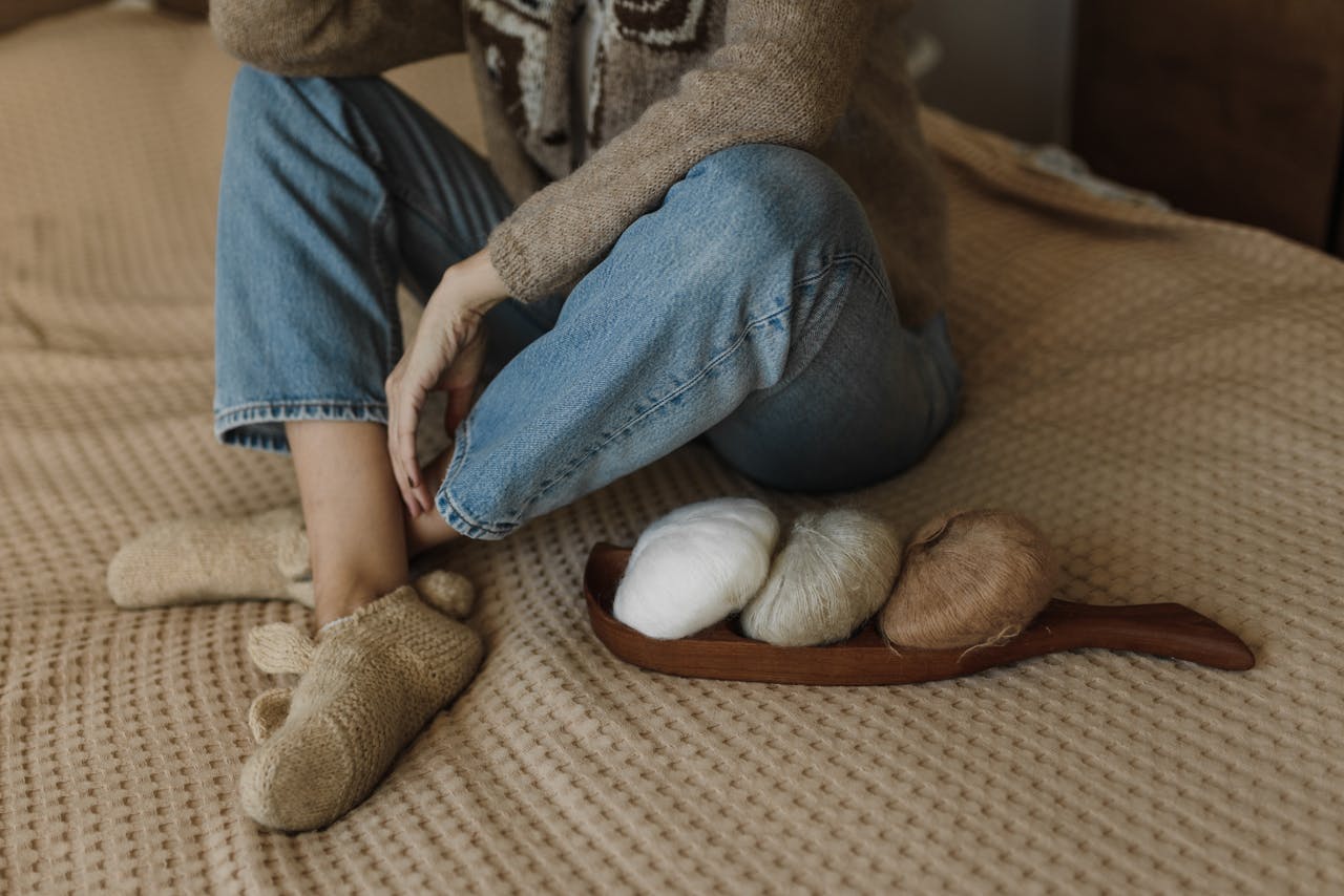 Woman sitting on a bed with yarn rolls, wearing cozy slippers, creating a warm knitting ambiance.